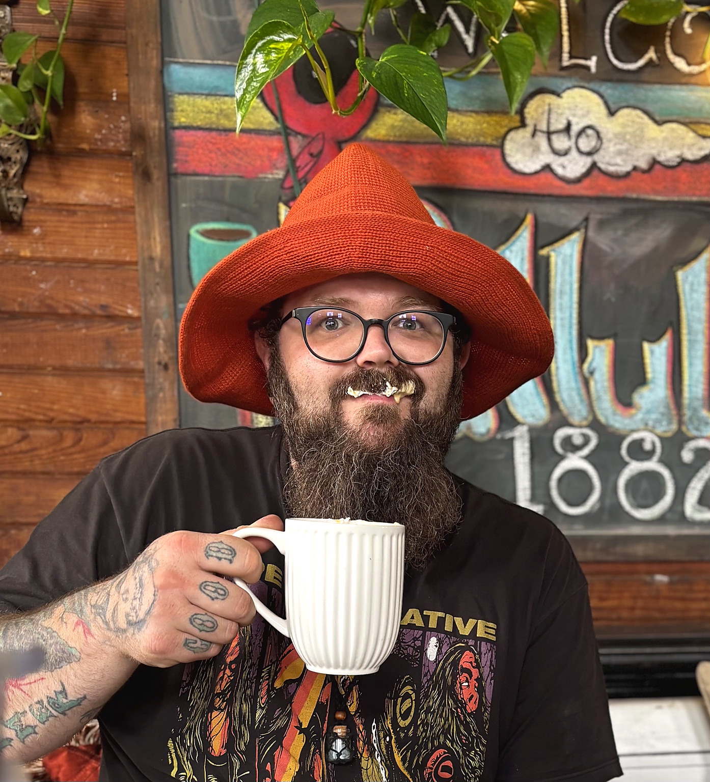 The maker behind Lace & Leather Arcane wearing a rust-red hat and glasses, smiling and holding a white coffee mug.
